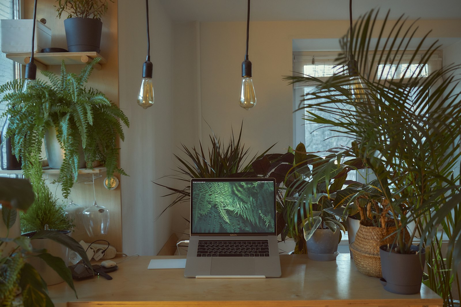 Mesa de trabalho com notebook e plantas, sob luminárias acesas